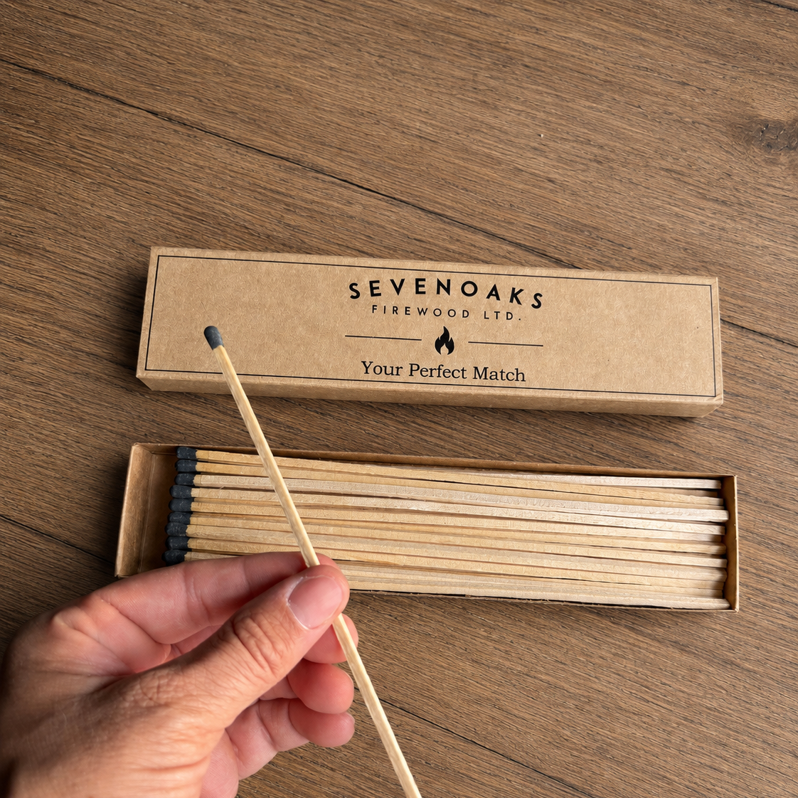 A hand holds a single match from a box of Sevenoaks Firewood Ltd. matches on a wooden tabletop.